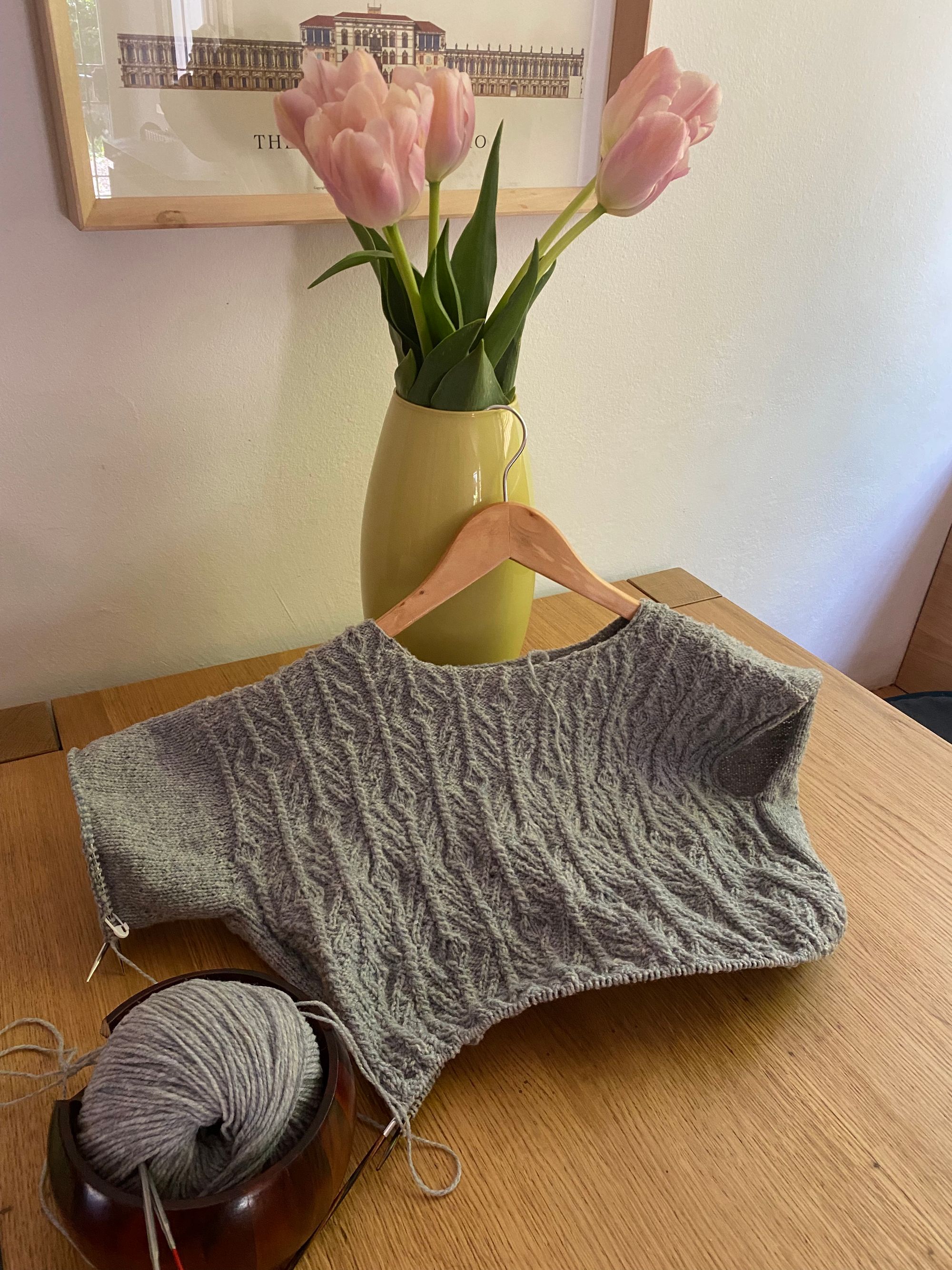 A yarn ball in a yarn holder, connected to a sweater in progress, hanging on a vase with tulips